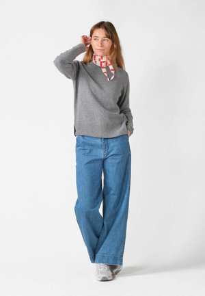 Vrouw met een grijze trui, wijde blauwe spijkerbroek, witte sneakers en een rood-wit geruit halsdoekje, staand met één hand in de zak.