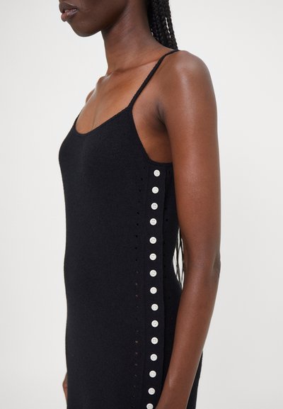 Schwarzes Strickkleid mit dünnen Trägern, das über eine seitliche Knopfschließung und kleine Ösen entlang des Saums verfügt. Glatte Textur.