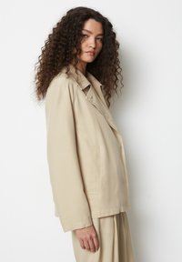 Beež puuvillane jakk klapiga kraega, küljetaskutega ja lõdva istuvusega, millel on sile tekstuur ja minimalistik disain.