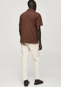 Camicia marrone a maniche corte con colletto, indossata con pantaloni bianchi slim-fit e sandali scuri. Tessuto liscio, fit rilassato e dettagli minimi.