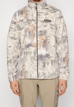 Mann trägt eine beige und graue Columbia Fleecejacke mit Waldmuster und durchgehendem Frontreißverschluss sowie beige Cargohosen.