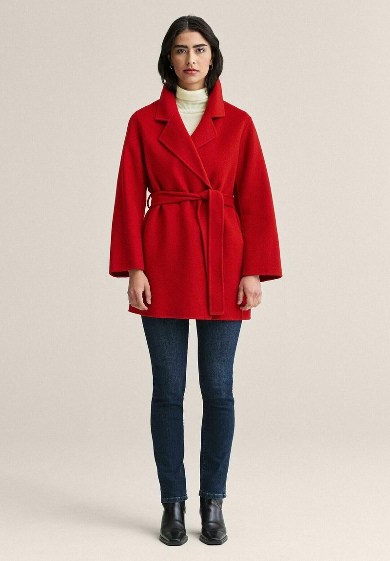 Punainen villakangastakki vyöllä, leveillä kauluksilla ja kolmen neljänneksen hihoilla, yhdistetty tummiin farkkuihin ja nilkkureihin. Yksinkertainen ja jäsennelty muotoilu.