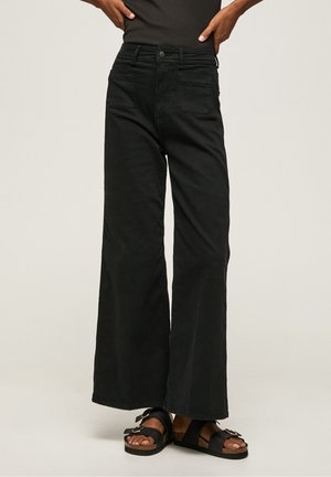 Personne portant un pantalon noir taille haute à jambes larges avec poches avant et des sandales noires à boucle, debout les mains sur les hanches.