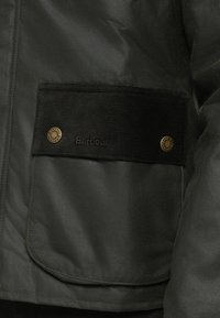 Μπουφάν puffer σε σκούρο πράσινο με τσέπη από κορντόι. Διαθέτει ορείχαλκινα κουμπώματα και ανάγλυφο λογότυπο "Barbour" στην τσέπη. Λεία υφή.