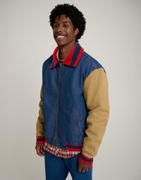 Τζιν και μπεζ μπουφάν bomber με κόκκινες ρίγες στον γιακά; περιλαμβάνει πλεκτές μανσέτες και στρίφωμα, καθώς και δύο πλαϊνές τσέπες, τοποθετημένο πάνω από πουκάμισο καρό.