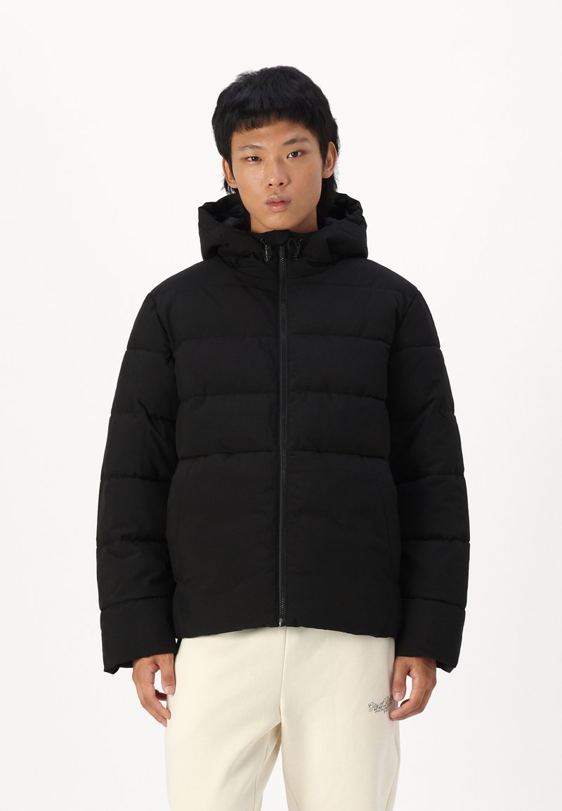 Sort pufferjakke med lynlås foran, brede horisontale sømme, hætte og lange ærmer, lavet af nylon. Båret med beige bukser.