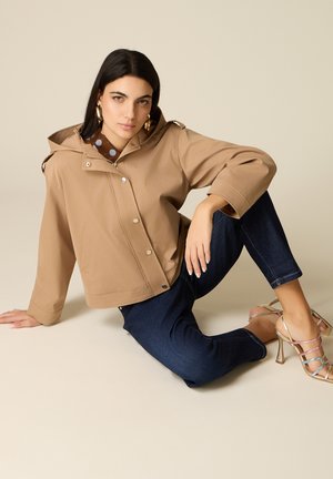 Donna con capelli scuri che indossa una giacca con cappuccio color sabbia, jeans blu scuro e sandali dorati con tacco alto, seduta su un pavimento beige contro uno sfondo semplice.