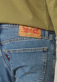 Paire de jeans en denim bleu avec poche arrière et étiquette en cuir marron clair portant un logo rouge et du texte, portés avec un haut vert olive.