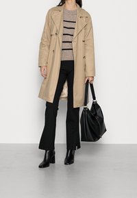 Beiger Trenchcoat mit doppelt geknöpften Knöpfen, gestreiftem braunem Pullover, schwarzen Schlaghosen und schwarzen Stiefeln mit Absatz. Schwarze Handtasche mit goldenen Akzenten.