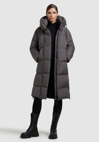Abrigo puffer largo gris con capucha, con un patrón acolchado, cierre frontal de cremallera y bolsillos laterales, combinado con botas de combate negras.