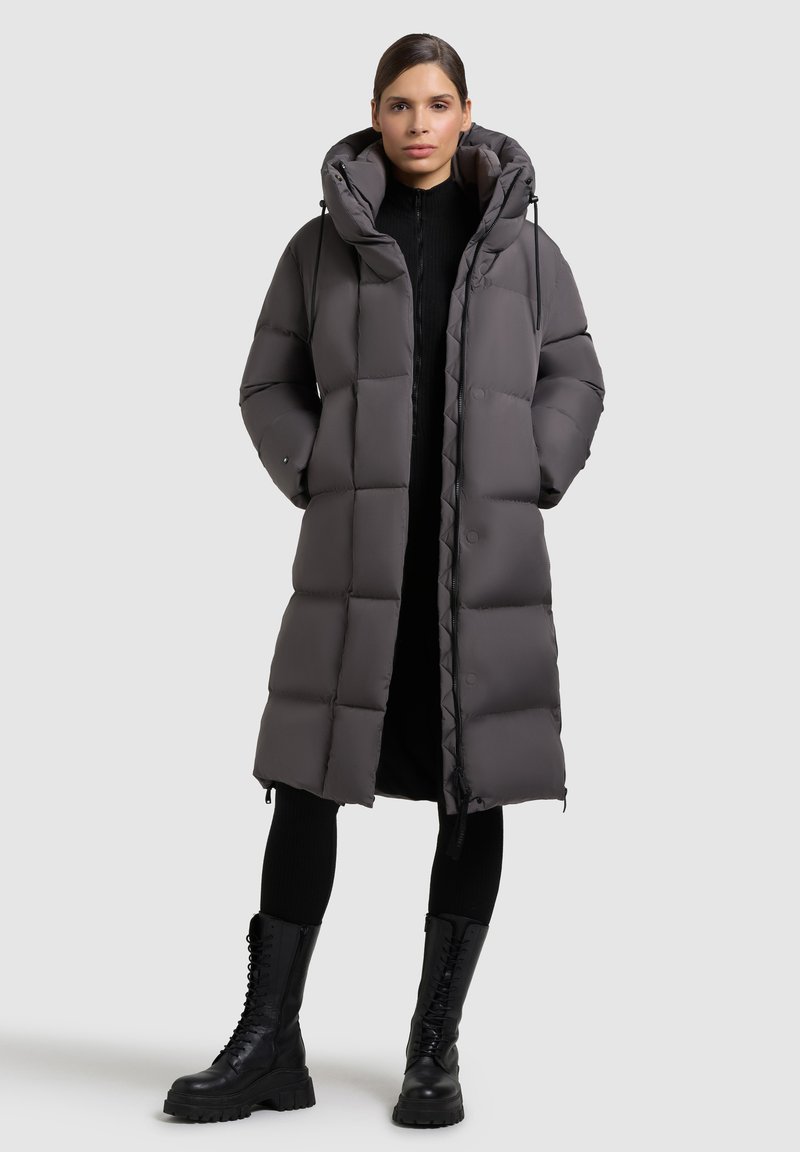 Abrigo puffer largo gris con capucha, con un patrón acolchado, cierre frontal de cremallera y bolsillos laterales, combinado con botas de combate negras.