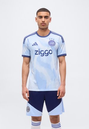 Ein junger Mann steht und trägt ein hellblaues und dunkelblaues Fußballtrikot mit Ziggo-Logo, Adidas-Streifen und Teamwappen auf Trikot und Shorts.