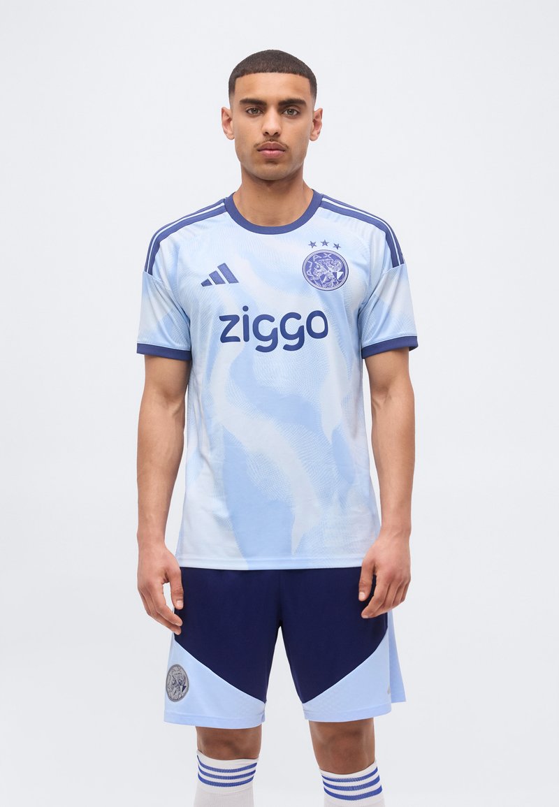 Un giovane uomo è in piedi indossando una divisa da calcio azzurro chiaro e blu navy con il logo Ziggo, le strisce Adidas e lo stemma della squadra su maglia e pantaloncini.