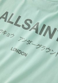 Mintgrøn T-shirt med fed sort tekst, der lyder "ALLSAINTS" samt yderligere tegn. Blødt stof med rund hals.