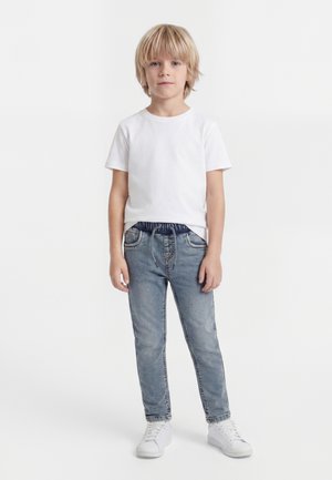 Băiat blond stând drept, purtând un tricou alb simplu, blugi albastru deschis cu snur și adidași albi, pe fundal alb.