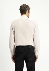 Camicia a maniche lunghe color beige chiaro con texture liscia, vestibilità su misura e polsini con bottoni visibili, abbinata a jeans neri scuri.