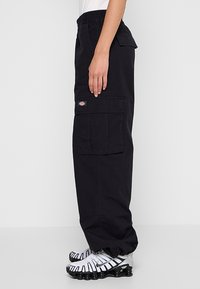 Personne portant un pantalon cargo noir avec des poches à rabats et des baskets blanches Nike Shox, debout devant un fond clair uni.