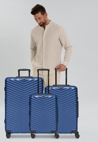 Homme en chemise beige tenant la poignée d'une valise rigide bleu moyen, avec deux valises similaires, plus grande et plus petite, à proximité.