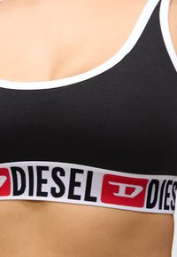 Soutien desportivo preto com detalhes brancos, apresentando uma larga faixa elástica com o logótipo "DIESEL" em cores vermelha e preta. Textura de tecido suave.