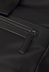 Tessuto in nylon nero con una superficie liscia, dotato di chiusura con zip e un tirante in pelle, e cuciture rinforzate lungo i bordi.
