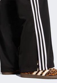 Czarne sportowe spodnie z białymi pionowymi paskami, z suwakiem u dołu, zestawione z brązowymi butami w panterkę.