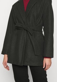Cappotto in lana grigio scuro con cintura, colletto a scialle, maniche lunghe e tasche laterali, indossato sopra un top rosso. Texture liscia, design minimalista.