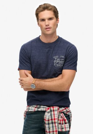 Navy blue short-sleeve t-shirt with a textured finish and printed logo on the chest. Light plaid flannel shirt tied around the waist.