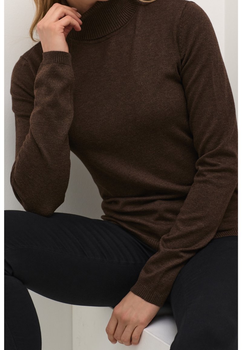 Personne portant un pull à col roulé marron et un pantalon noir, assise avec une main posée sur une surface blanche et l'autre près du visage.