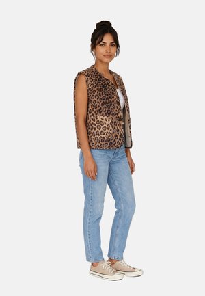 Donna che indossa un gilet senza maniche con stampa leopardata, top bianco, jeans azzurri chiari e sneaker beige, in piedi su sfondo bianco.
