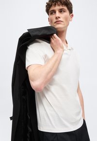 Νεαρός άνδρας που φοράει λευκό πόλο μπλουζάκι και μαύρο παντελόνι, κρατώντας μαύρο σακάκι στον ώμο του, κοιτάζει στο πλάι μπροστά σε ένα απλό φόντο.