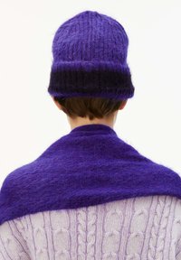 Person, die eine lila und schwarze Strickmütze und einen passenden flauschigen lila Schal über einem hell-lila Zopfmusterpullover trägt, von hinten gesehen.