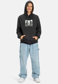 Čierna kapucňová mikina s veľkým strieborným logom DC na prednej strane, v kombinácii so sivými voľnými džínsami a bielymi teniskami. Neformálny dizajn.