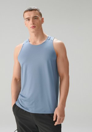 Junger Mann mit kurzem Haar, bekleidet mit einem hellblauen, ärmellosen Sporttop und schwarzen Hosen, vor einem schlichten grauen Hintergrund stehend.