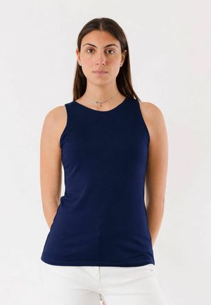 Donna con lunghi capelli scuri che indossa una canottiera blu navy e pantaloni bianchi, in piedi contro uno sfondo bianco uniforme.