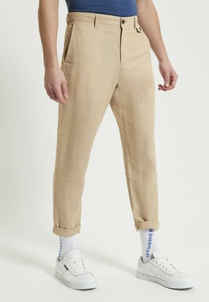 Pantaloni beige in misto lino con vestibilità affusolata, dotati di tasche laterali e risvolti, abbinati a sneakers bianche e calze a righe.