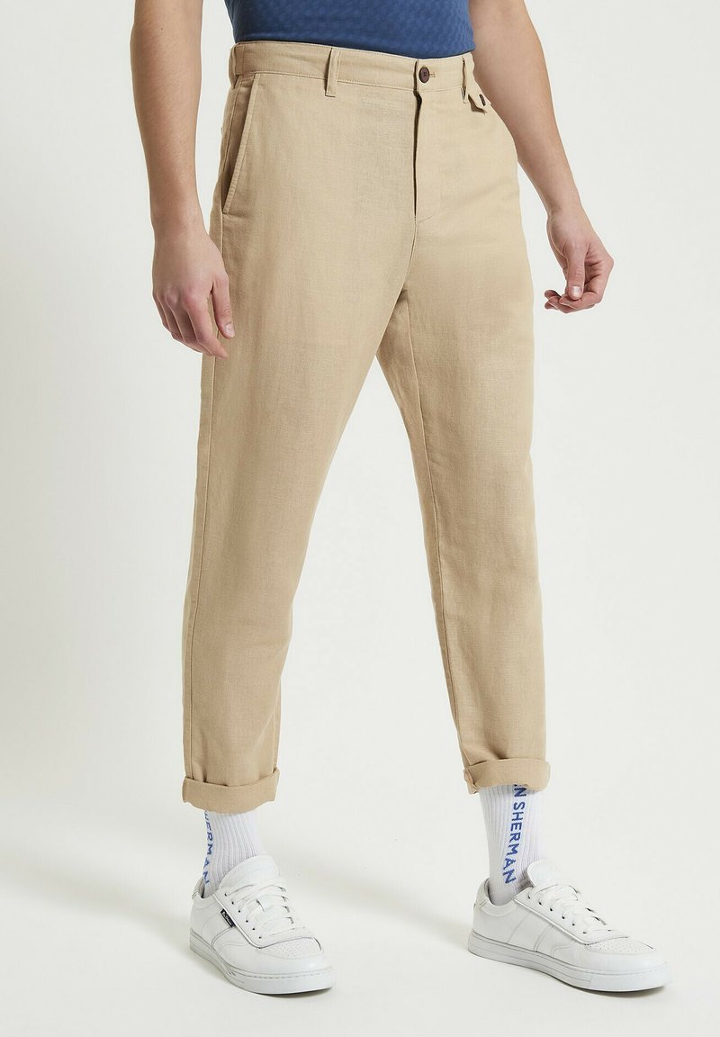 Pantaloni beige in misto lino con vestibilità affusolata, dotati di tasche laterali e risvolti, abbinati a sneakers bianche e calze a righe.