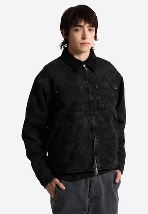 Jong persoon met halflang donker haar draagt een zwarte jas met rits en handzakken en een grijze broek, staand tegen een effen achtergrond.