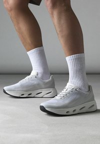 Bílé sportovní boty s síťovinovým svrškem, šedými akcenty a silnou podešví s výřezovými detaily. V kombinaci s ribbovanými bílými ponožkami.