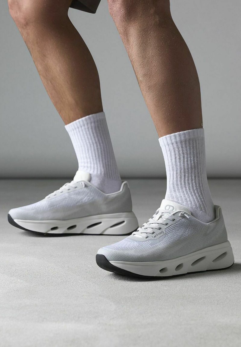 Bílé sportovní boty s síťovinovým svrškem, šedými akcenty a silnou podešví s výřezovými detaily. V kombinaci s ribbovanými bílými ponožkami.