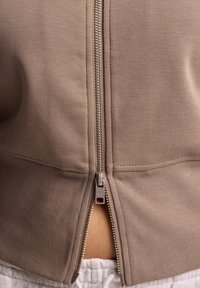 Felpa con zip marrone, con un collo alto e dettaglio della zip visibile sulla schiena, realizzata in tessuto testurizzato. Finiture delle cuciture curate.