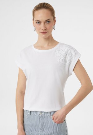T-shirt en coton blanc avec des manches courtes à revers, ornée d'un motif de broderie florale sur l'épaule gauche. Coupe simple et décontractée.