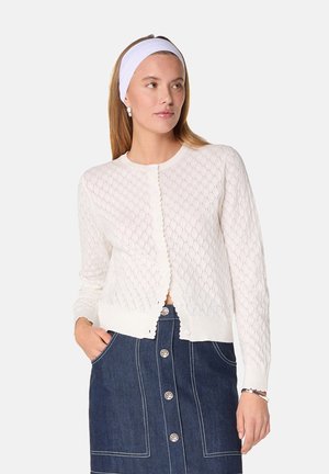 Cardigan tricoté blanc avec un motif en losange, col rond, bords festonnés et fermeture à boutons, associé à une jupe en denim.