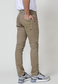 Beige slim-fit nadrág hátsó zseb részlettel, sima textúrával. Szürke és kék sportcipővel párosítva.