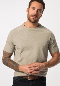 Svetle šedé tričko s krátkým rukávem a kulatým výstřihem, vyrobené z měkké tkaniny. Částečně viditelná je tetovaná ruka modelu.