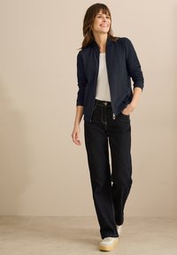 Veste bombardier marine à motif texturé, t-shirt blanc et jeans noirs taille haute. Le modèle porte des baskets blanches avec une semelle beige.