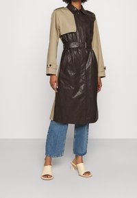 Płaszcz trench z beżową górą i ciemnobrązowym skórzanym dołem, z paskiem w talii, guzikami przy mankietach oraz pojedynczym detalem klapy na jednym ramieniu przy kołnierzu.