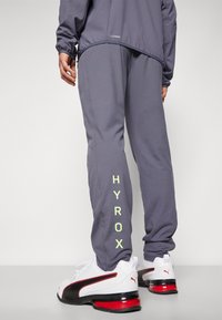 Grijze sportbroek met "HYROX" in neon groen gedrukt. Gecombineerd met witte en zwarte sneakers met rode accenten. Soepele textuur en slim fit.