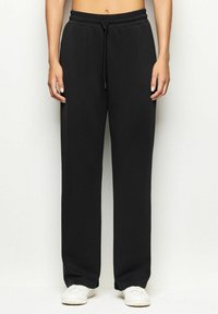 Persona che indossa pantaloni tuta neri con coulisse e sneakers bianche, in piedi contro uno sfondo chiaro e semplice.