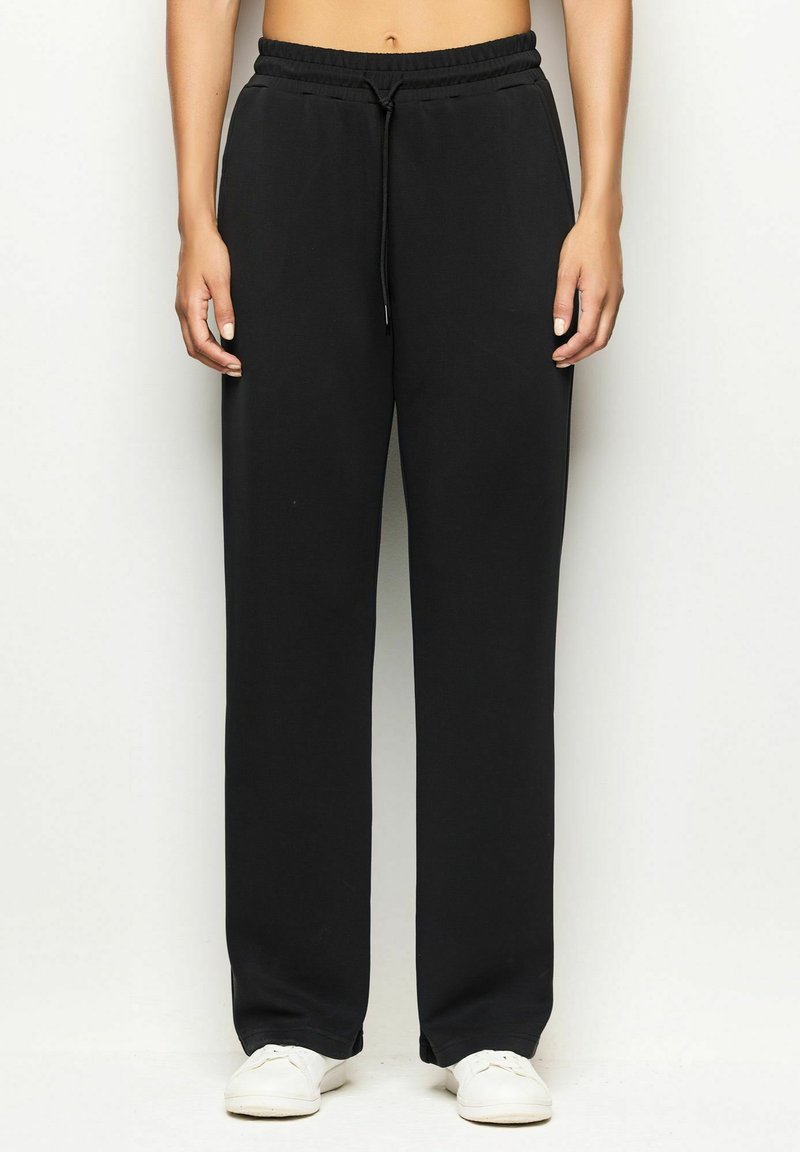 Persona che indossa pantaloni tuta neri con coulisse e sneakers bianche, in piedi contro uno sfondo chiaro e semplice.