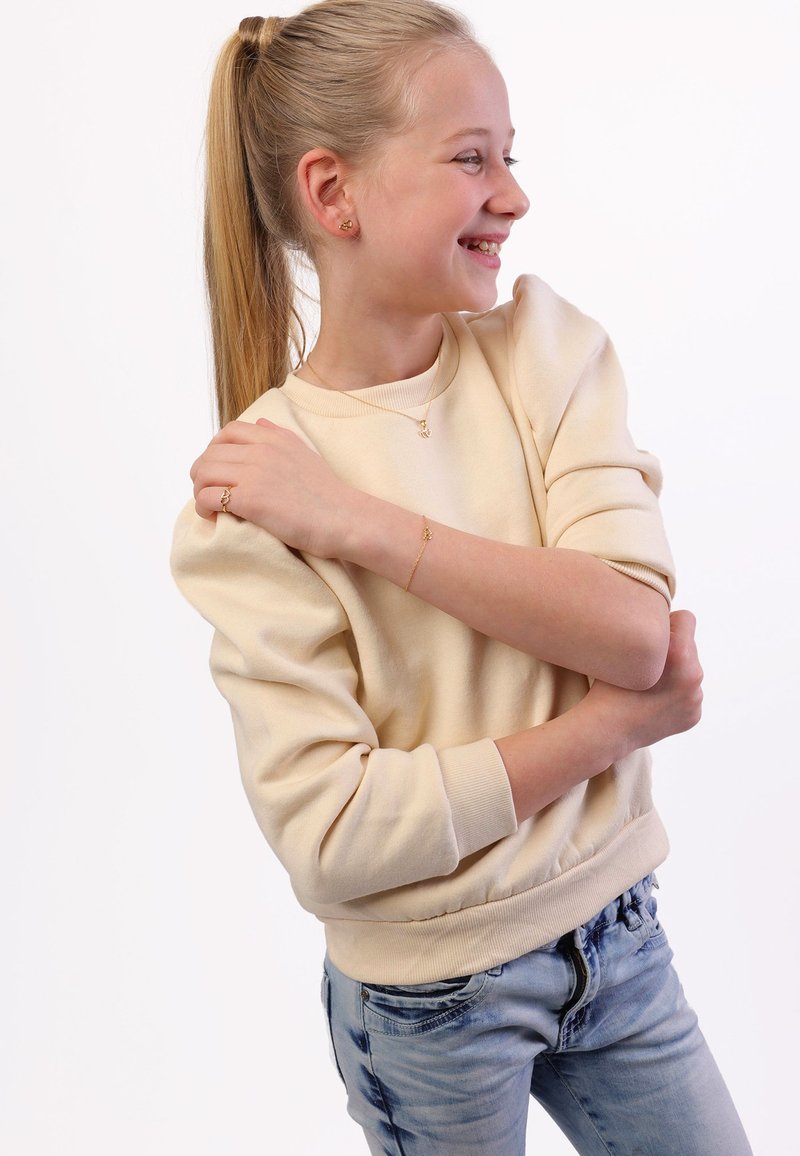 Beige sweatshirt met poffige mouwen, geribbelde manchetten en tailleband, gecombineerd met lichtblauwe jeans. Sieraden bestaan uit een ketting, armband en ring.